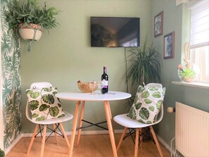 Ein Zimmer mit einem runden Tisch, zwei Stühlen mit Blattmuster, einer Weinflasche, einem Korb und einem Fernseher an der Wand.
