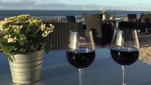 Twee wijnglazen op een tafel met uitzicht op zee, een bloempot en een schaal met noten.