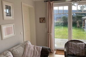 Sala de estar com sofá, cadeira de vime, porta de vidro para um jardim e quadros nas paredes.