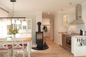 A modern kitchen with a central wood-burning stove, white cabinetry, and a dining area with a wooden table and chairs. - Holiday apartment with 2 bedrooms (Wangerooge)