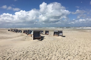 Ein Strand mit Reihen von gestreiften Liegestühlen und Sonnenschirmen, ein sandiger Uferbereich und das Meer im Hintergrund.