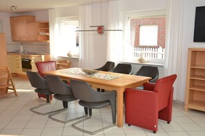 Een eetgedeelte met een houten tafel en stoelen, een keuken op de achtergrond, en een raam met uitzicht op een gebouw van baksteen.
