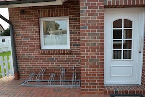 A brick building with a white door and window, a metal bike rack, and a white picket fence in the background. - MeerZeit - Süderpfad 2b (Borkum)