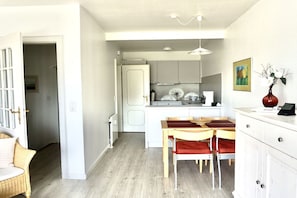 Cocina compacta con armarios blancos, una zona de comedor con mesa y sillas de madera, y una lámpara colgante.