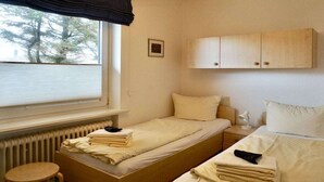Ein Zimmer mit zwei Betten, einem Fenster mit Jalousien, einem Heizkörper, einem kleinen Tisch und einem Schrank.