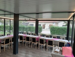 Un restaurant amb una taula llarga preparada per dinar, grans finestres i una vista cap a l'exterior.