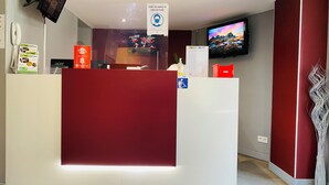 A hotel reception desk with a red counter, a mounted television displaying a scenic mountain view, and a sign indicating a mandatory mask policy. - Appart'hôtel Martinez (Grenay)