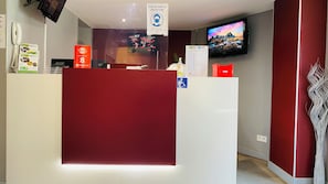 A hotel reception desk with a red counter, a mounted television displaying a scenic mountain view, and a sign indicating a mandatory mask policy. - Appart'hôtel Martinez (Grenay)