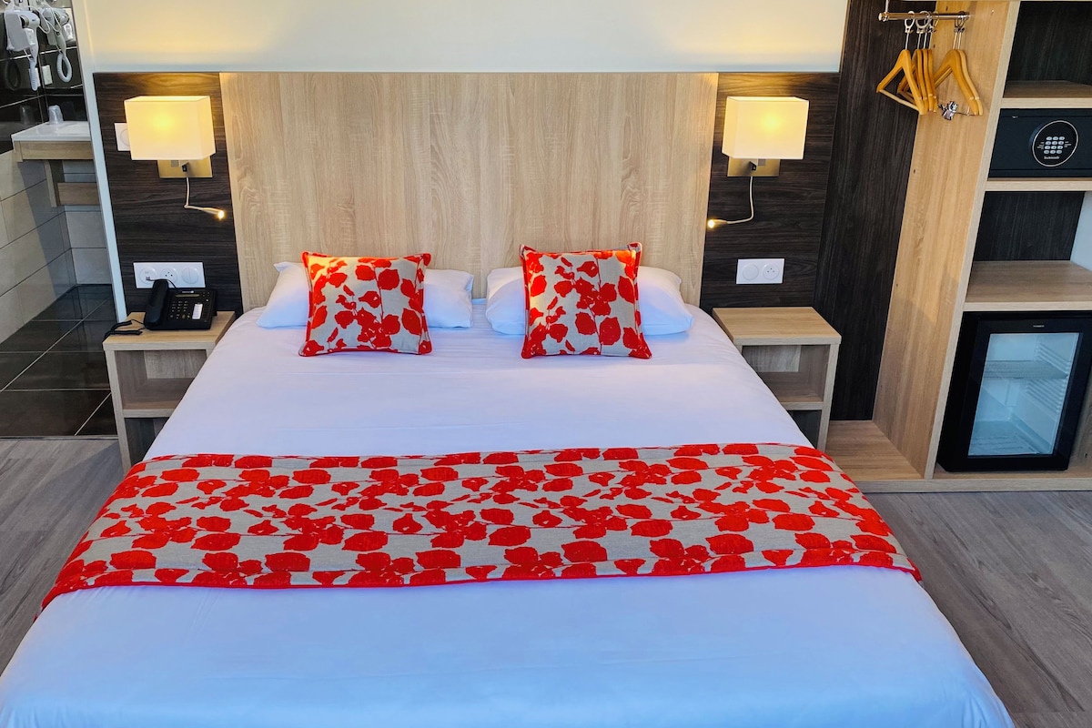 A hotel room with a large bed, two bedside tables, a nightstand, a built-in mini-fridge, and a wall-mounted lamp.