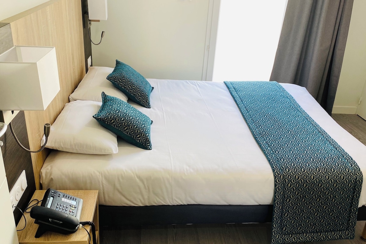 A hotel room with a bed, pillows, a bedside table with a phone, and a lamp.