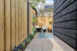 En smal udendørs terrasse med et træhegn, et lille bord med stole og en blå, mønstret måtte.