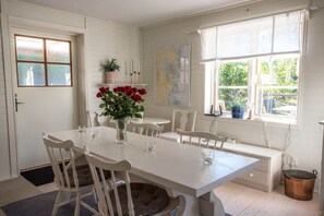 A dining area with a white table and chairs, a vase with red flowers, and a window with a view of greenery.