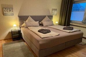 Cama doble con dos almohadas y una colcha, mesita de noche con lámpara y cuadros en las paredes.