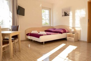 Un dormitorio con cama, escritorio, silla, mesita de noche, televisión, ventana y un cuadro en la pared.
