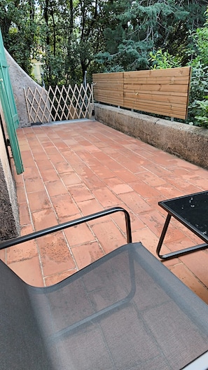 En uteplats med terrakottafärgat klinkergolv, ett trästaket och ett metallbord.
