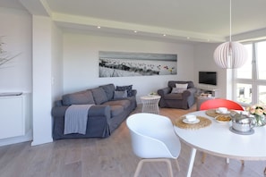 Ein modernes Wohnzimmer mit einem grauen Sofa, einem weißen Sessel, einem kleinen runden Tisch und einem Strandbild an der Wand.
