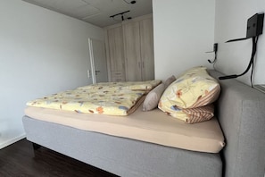 A single bed with floral bedding, a grey sofa, and a wardrobe in a room with white walls and a wooden floor. - Gertrude's Country House (Juist)