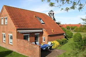 Ein Backsteinhaus mit einem geneigten, rot gedeckten Dach, einem kleinen Anbau mit Schornstein und einer Terrasse mit Tisch und Stühlen.