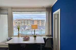 Habitación de hotel con una mesa de comedor, sillas, una lámpara, una planta en maceta y vistas de edificios a través de un ventanal grande.