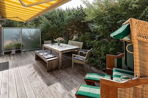 An outdoor patio with a wooden table and chairs, a bench, and a wicker chair with cushions. - The Captain's House by the Sea (Norderney)
