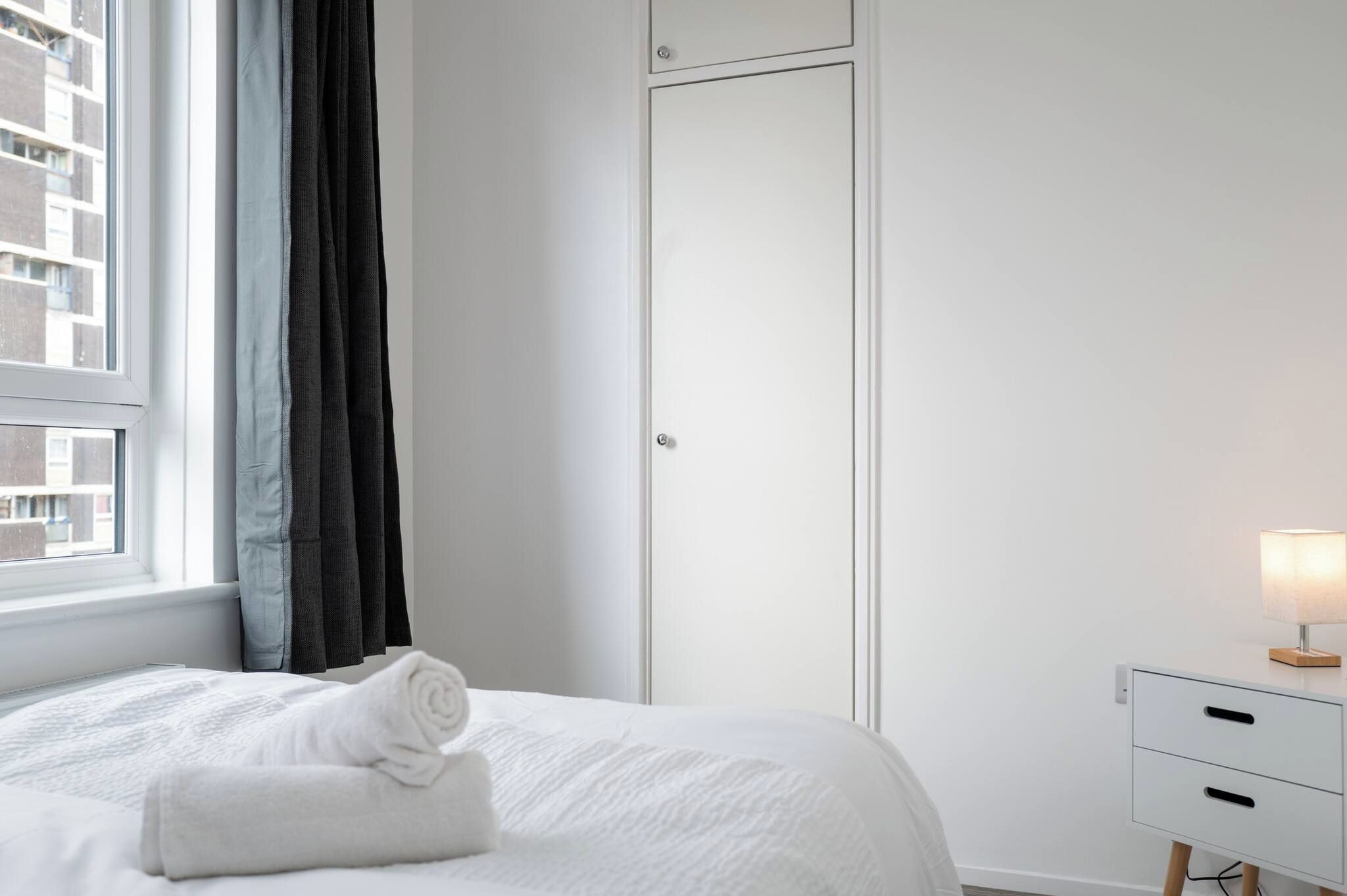 A bedroom with a bed, a window with curtains, a wardrobe, and a bedside table with a lamp.