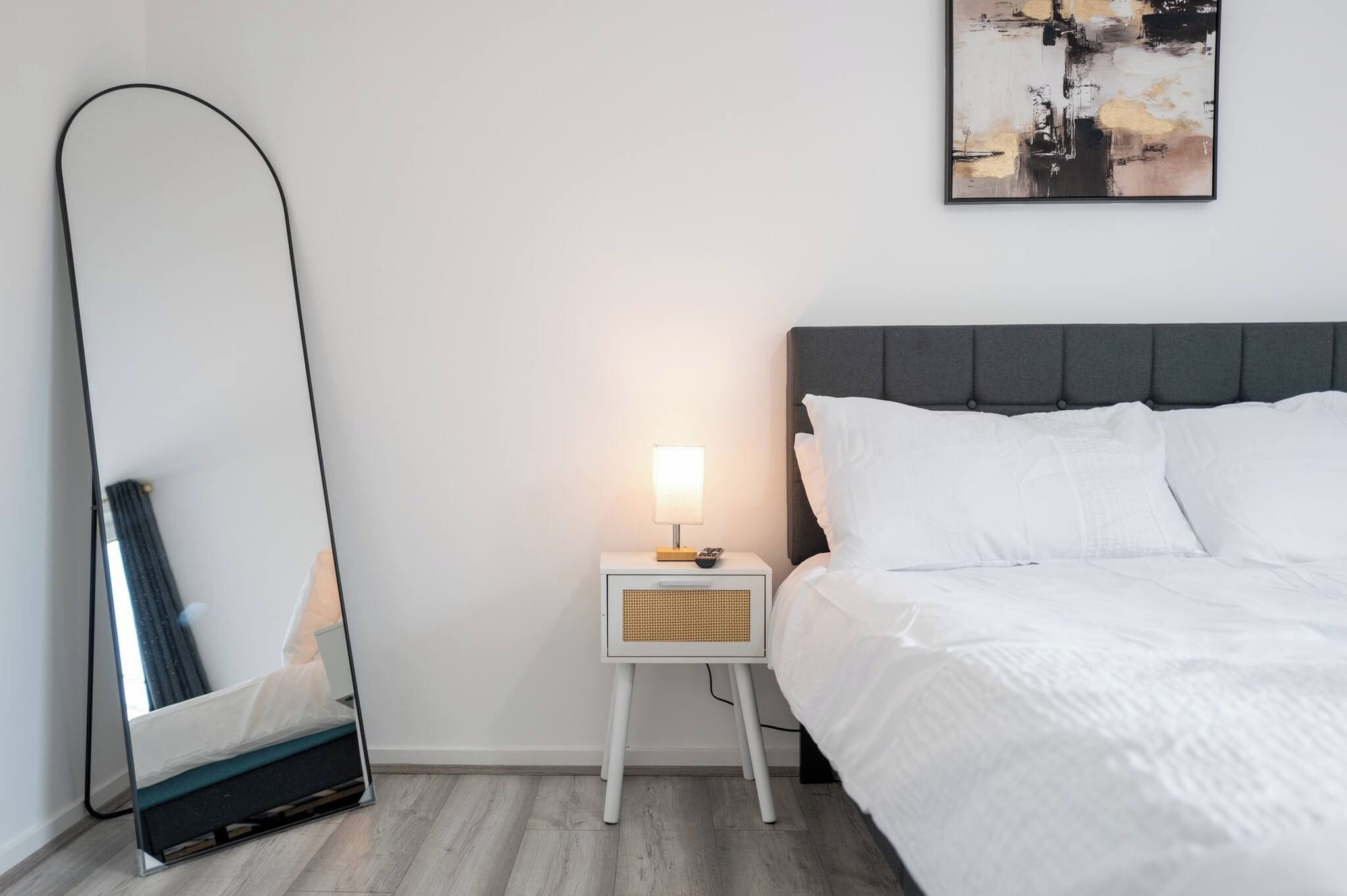 A bedroom with a bed, a nightstand with a lamp, a mirror, and a painting on the wall.
