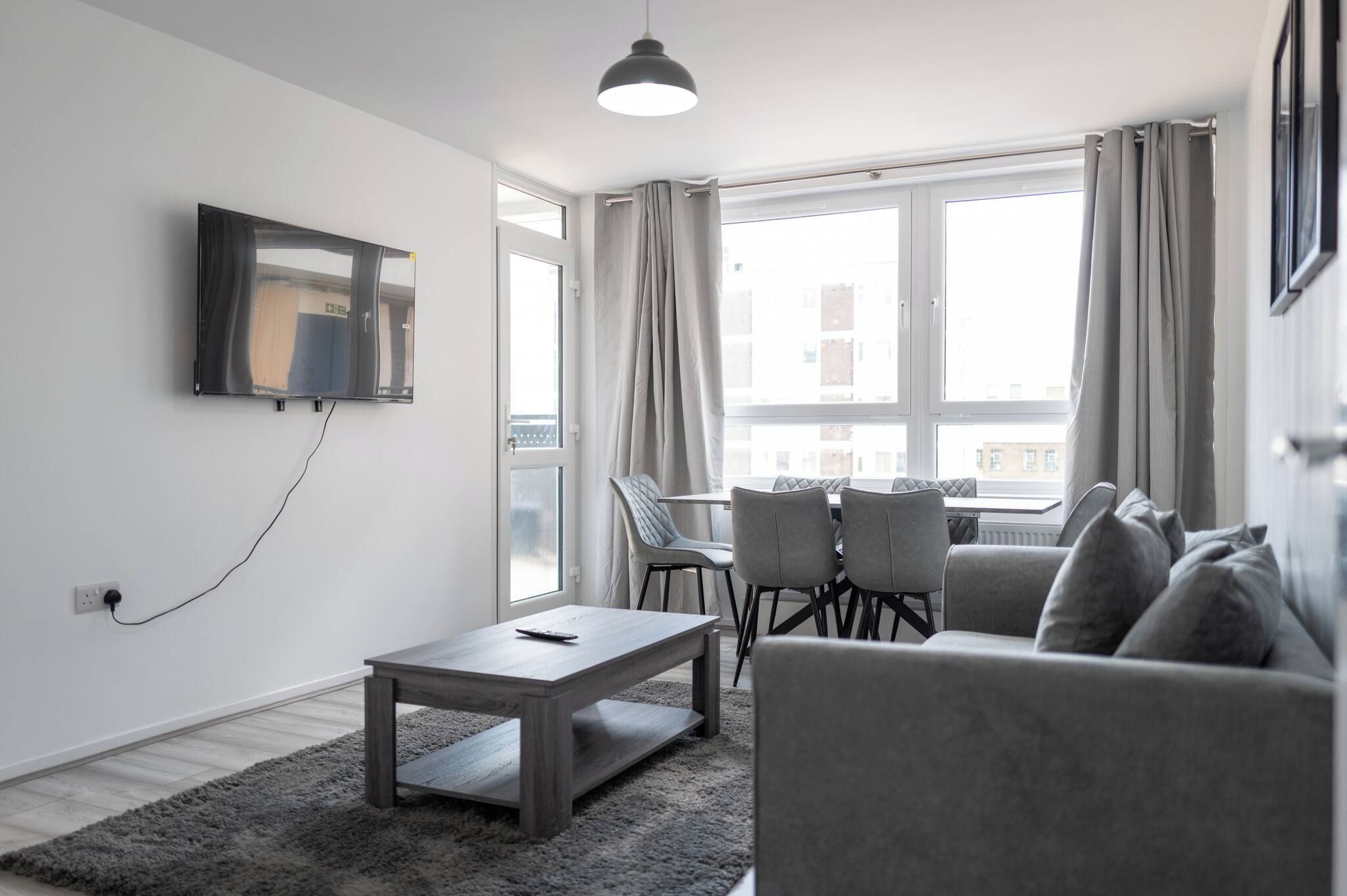 A modern living room with a flat-screen TV mounted on the wall, a grey sofa, a coffee table, and a dining area with chairs.