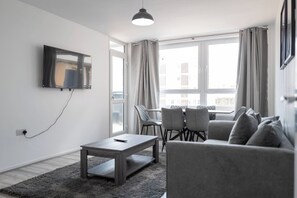 Ein modernes Wohnzimmer mit einem an der Wand befestigten Flachbildfernseher, einem grauen Sofa, einem Couchtisch und einem Essbereich mit Stühlen.