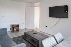 A modern living room with a grey sofa, a coffee table, a small wooden side table, and a television mounted on the wall. - Modern 3BD Flat Hoxton & Shoreditch N1 (London)