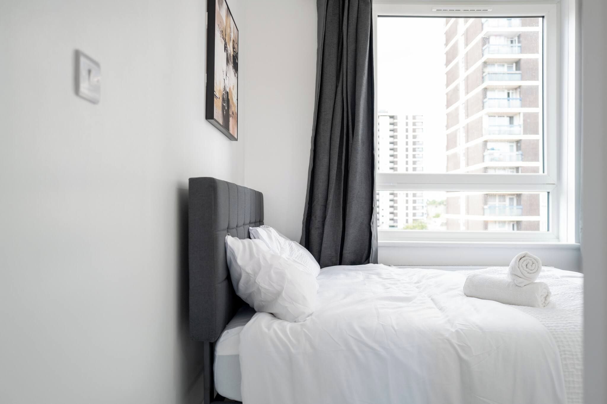 A hotel room with a bed, white bedding, a dark headboard, a window with a view of buildings, and a rolled towel on the bed.