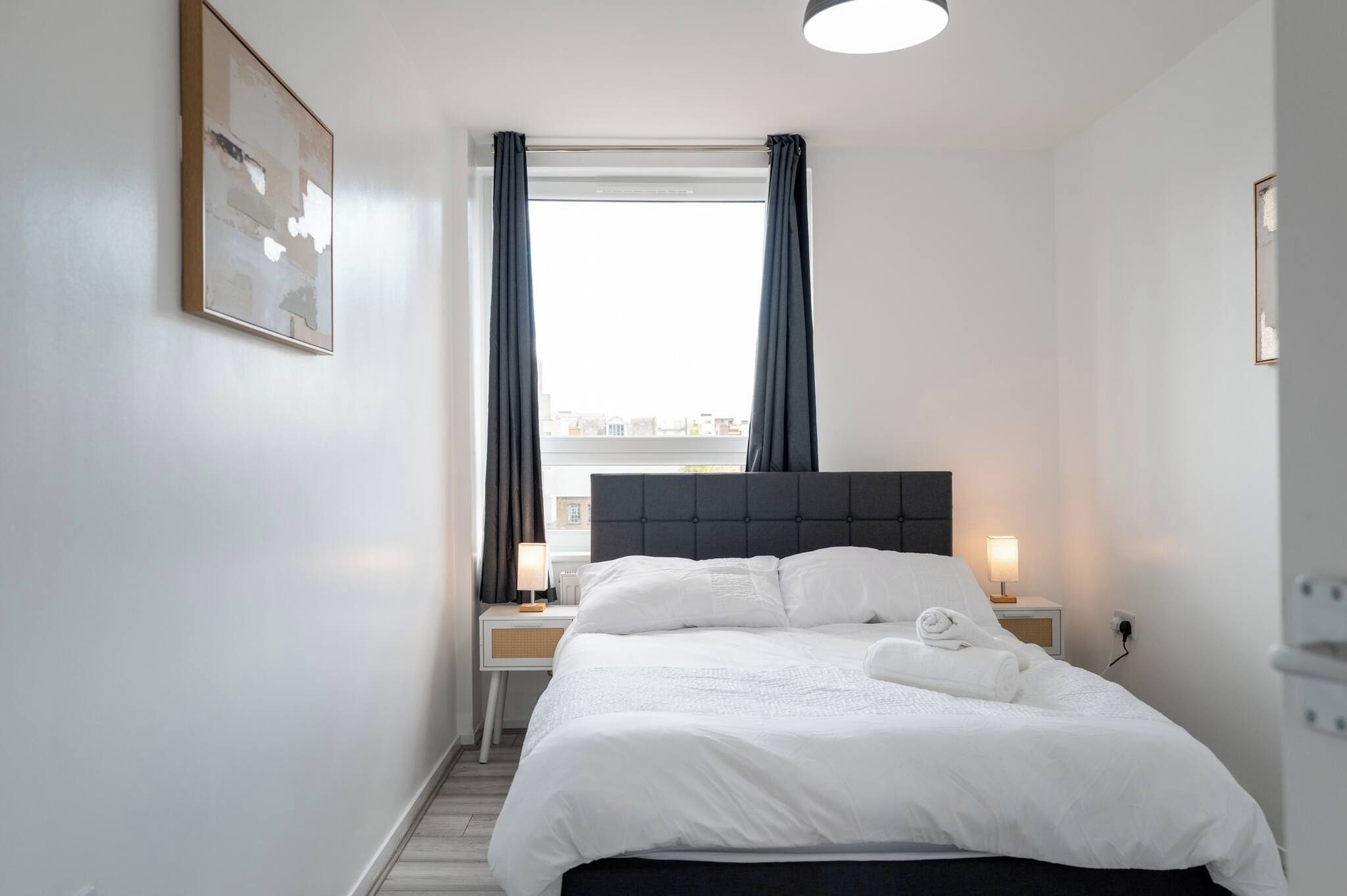A hotel room with a bed, bedside tables, a window with curtains, and a view of the city.