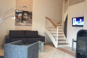 A living room with a sofa, a glass coffee table, a modern floor lamp, a staircase, and a television mounted on the wall. - Holiday apartment with 2 bedrooms (Wendisch Rietz)