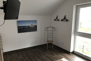 Ein Zimmer mit Holzboden, einem einzelnen Sessel, einem an der Wand befestigten Fernseher und einem Fenster mit Blick ins Freie.