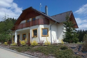 Ein zweigeschossiges Haus mit Balkon, Schornstein und einem Garten mit gelben Blumen.