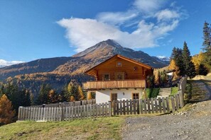 Eine Holzhütte mit Balkon, umgeben von einem Holzzaun, vor einer Berg- und Laublandschaft.