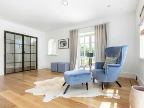 A spacious room with a large window, a blue armchair, a small table, and a white fur rug. - Holiday Home Pidder Hus Kampen / Sylt (Kampen)