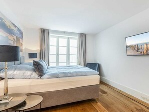 A bedroom with a bed, a bedside table, a lamp, a window with curtains, and a wall-mounted picture. - Holiday Home Pidder Hus Kampen / Sylt (Kampen)