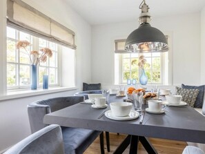 A dining area with a table set for four, surrounded by chairs, with a large window providing natural light. - Holiday Home Pidder Hus Kampen / Sylt (Kampen)