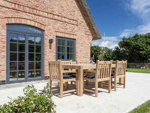 A brick building with a large window and a door, a patio with a table and chairs, and a garden with plants. - Holiday Home Pidder Hus Kampen / Sylt (Kampen)