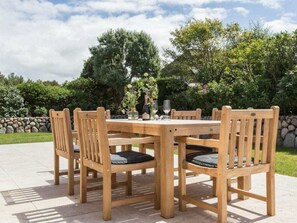 A wooden outdoor dining table with six chairs, set on a patio with a stone wall and greenery in the background. - Holiday Home Pidder Hus Kampen / Sylt (Kampen)