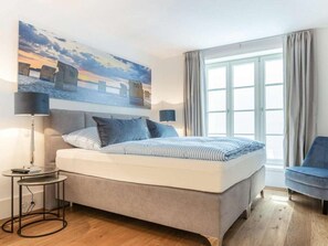 A bedroom with a bed, a bedside table, a chair, a window, and a painting on the wall. - Holiday Home Pidder Hus Kampen / Sylt (Kampen)