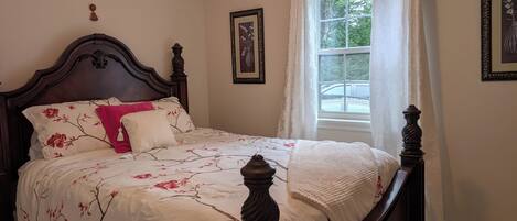 A bedroom with a wooden bed frame, floral bedding, a window with curtains, and a framed picture on the wall.