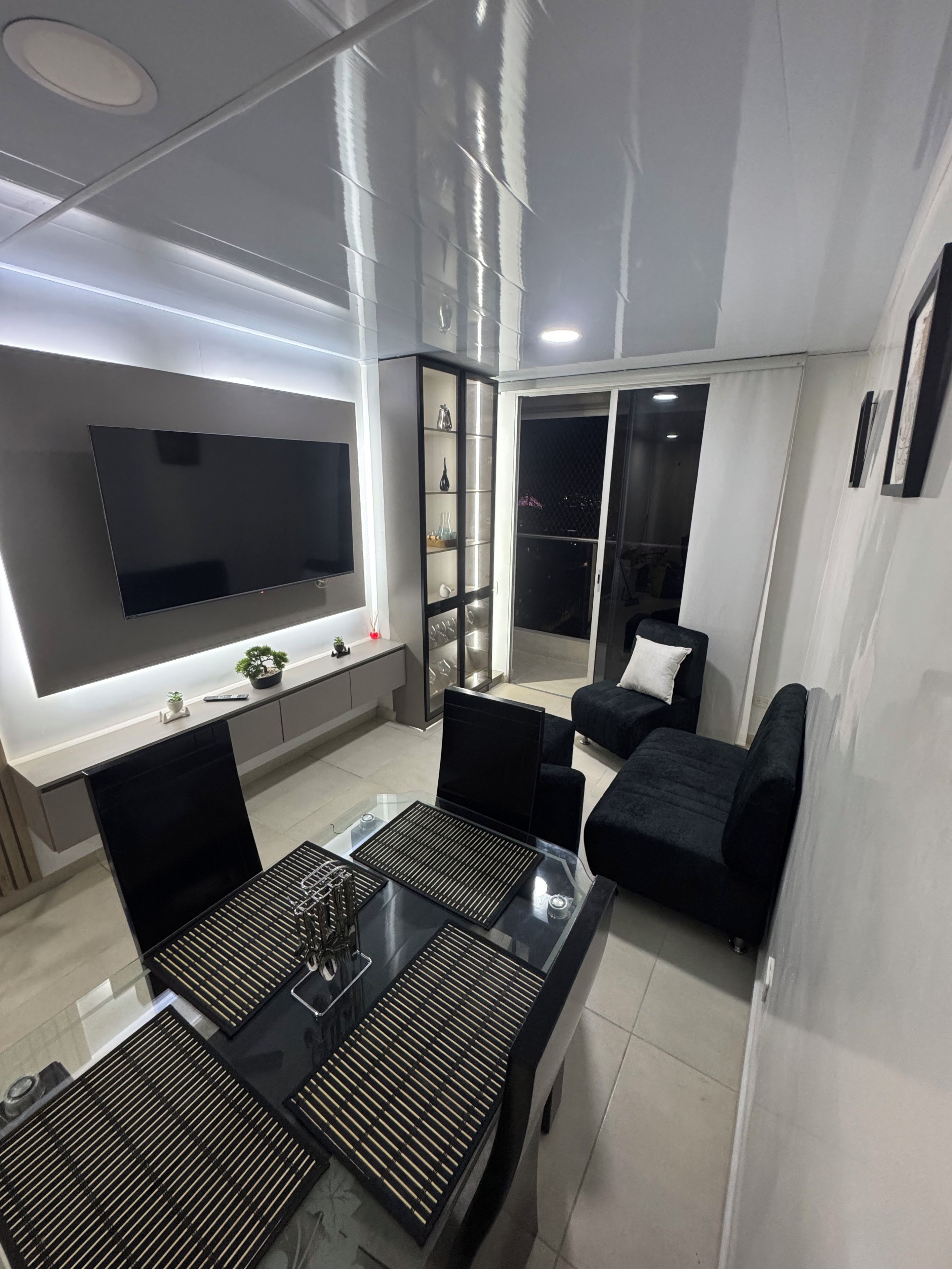 A modern living room with a flat-screen TV, black sofas, a glass coffee table, and a sliding glass door leading to a balcony.