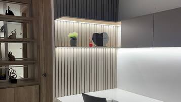 A modern office space with a white desk, a grey office chair, a shelving unit with decorative items, and a wall-mounted light panel.