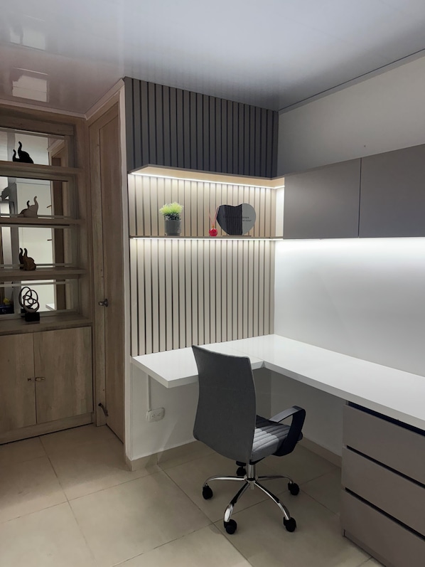 Un espai d'oficina modern amb escriptori blanc, cadira de color gris, una prestatgeria amb objectes decoratius i un plafó d'il·luminació penjat a la paret.