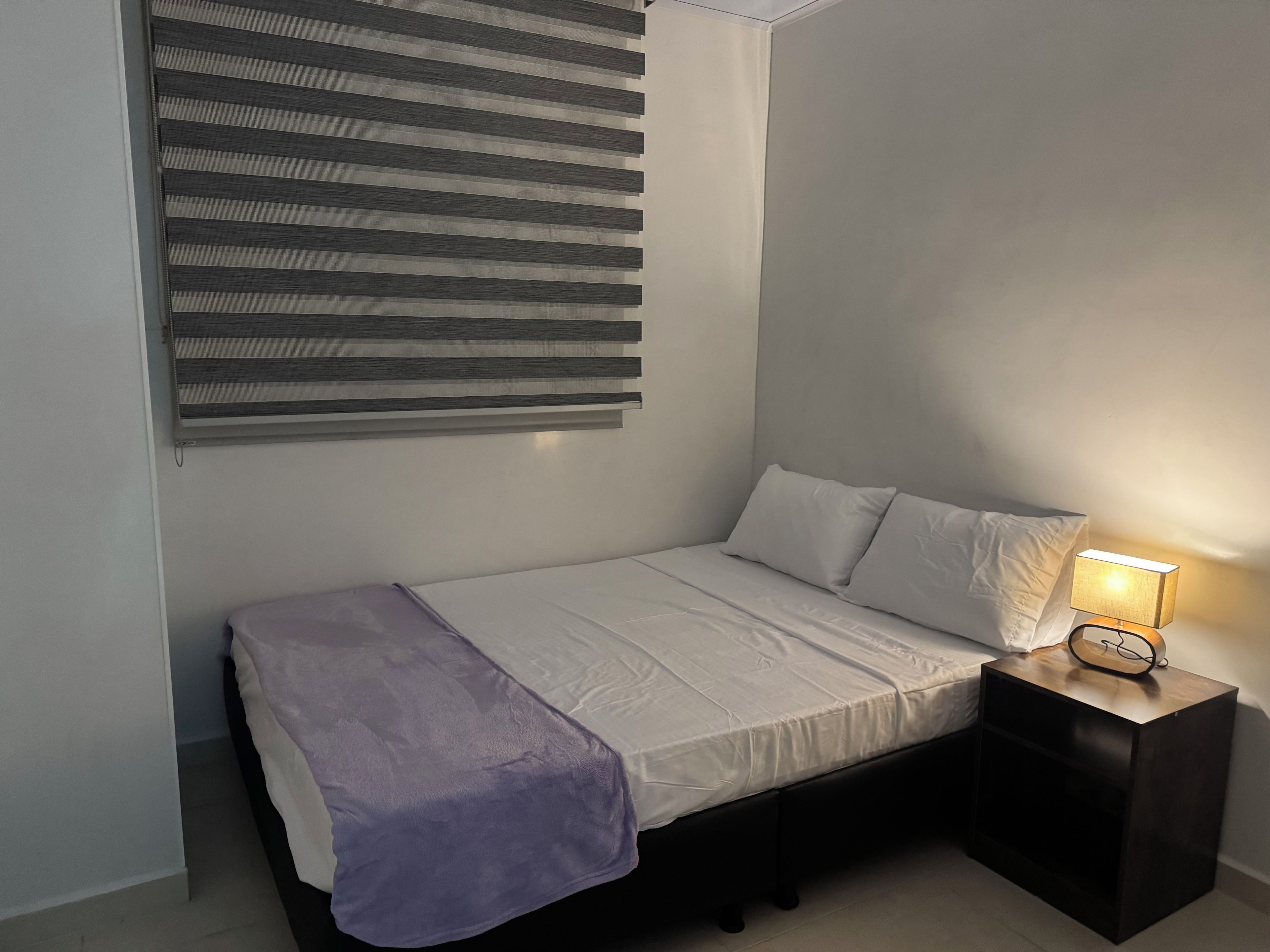 A single bed with a white mattress and pillows, a bedside table with a lamp, and a window with horizontal blinds.