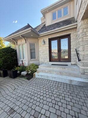 Une maison à deux étages avec une façade en pierre, une porte d’entrée dotée de panneaux de verre et un perron pavé de briques.