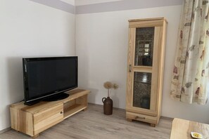 A room with a flat-screen TV on a wooden stand, a tall cabinet with glass doors, and a vase with dried flowers on the floor. - Holiday apartment with 2 bedrooms (Straupitz)