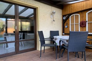 Eine Terrasse im Freien mit Tisch und Stühlen, eine Glastür zu einem überdachten Bereich und eine Holzverkleidung mit einer Laterne.