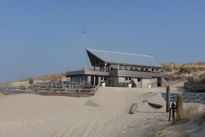 A building with a sloped roof, situated on a sandy beach with a wooden fence in the foreground. - Bredene Fisherman's House (Bredene)